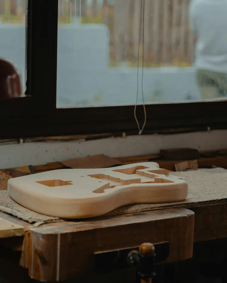 Pieza de madera desbastada en forma de cuerpo de guitarra, apoyada sobre el banco junto a la ventana del taller